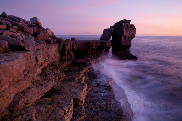 A large area of angular rock balances on the edge of a sheer cliff above the sea. The backdrop is a purple/pink sunset image. 
